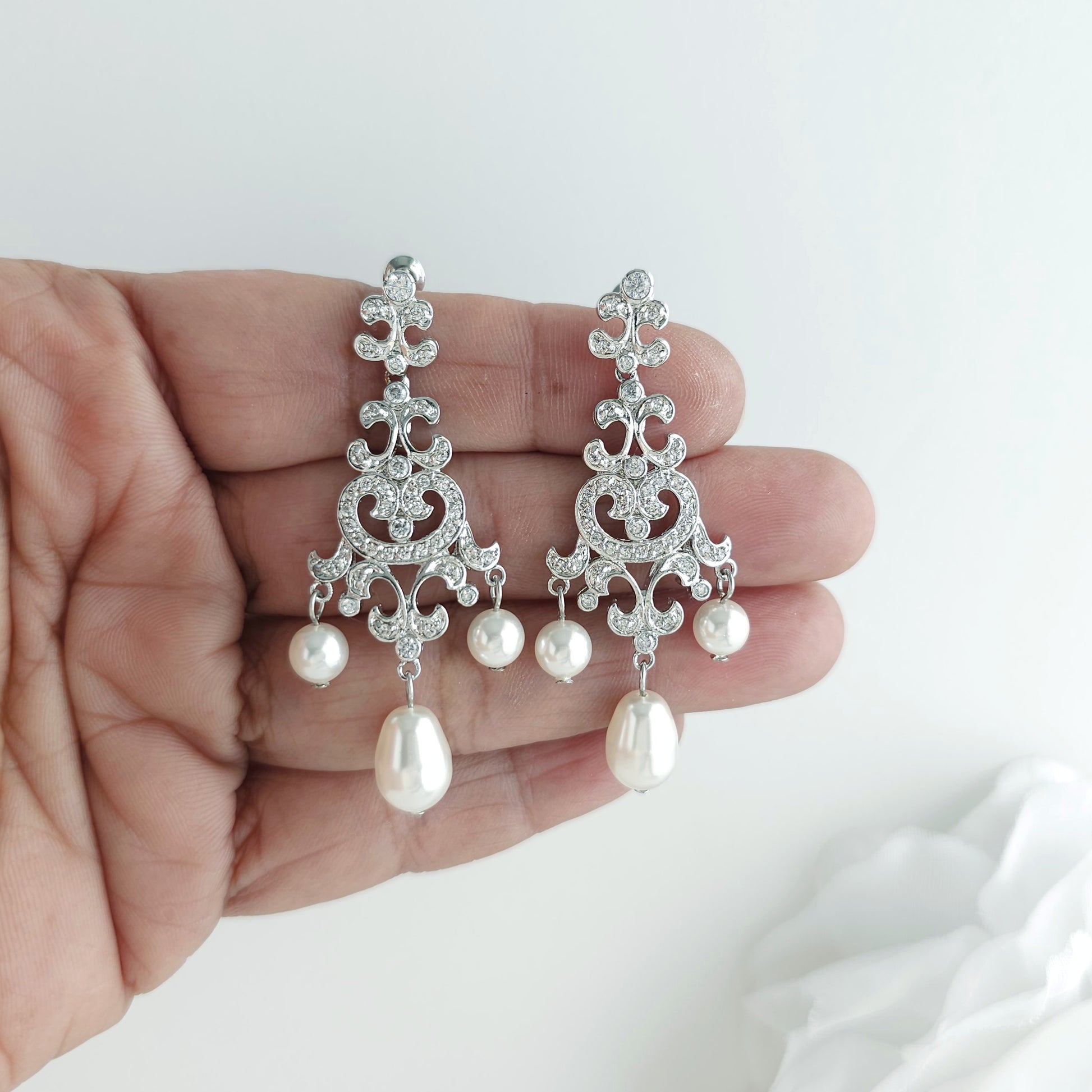A pair of ornate silver-toned dangle earrings with clear crystal filigree and white pearl drops, held in a person's hand against a soft white background to showcase the size of the earring- Lightweight silver vintage style pearl drop chandelier earrings featuring cubic zirconia and flawless round crystal pearls.