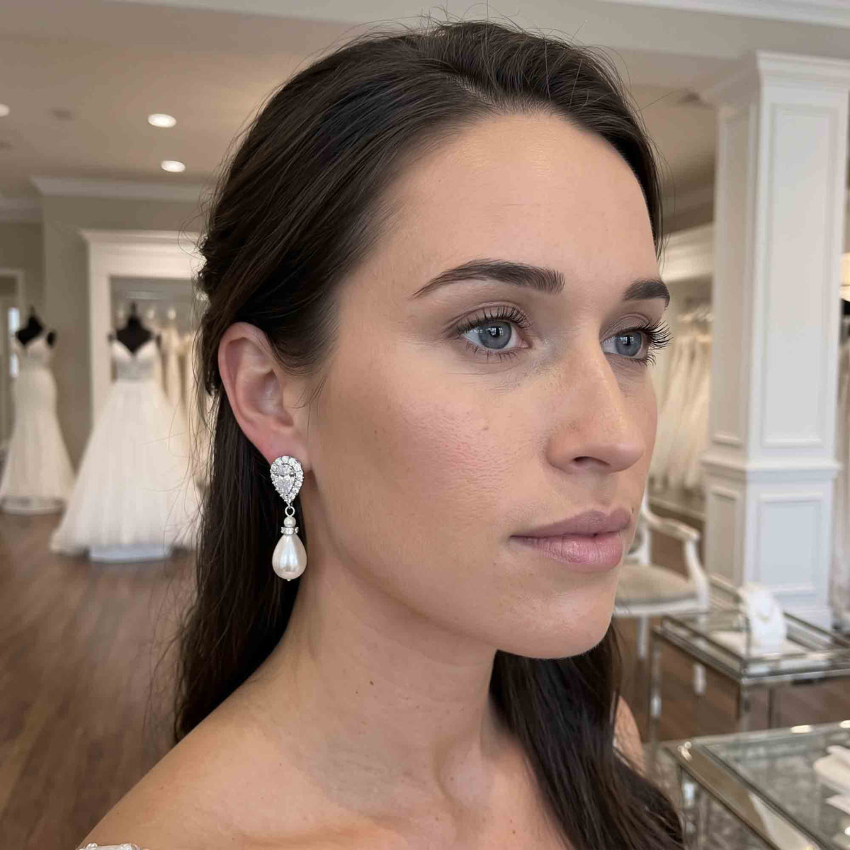 A woman wearing a dangling teardrop pearl earring with a sparkling crystal heart-shaped top, photographed inside a bridal shop with white wedding dresses visible in the background.