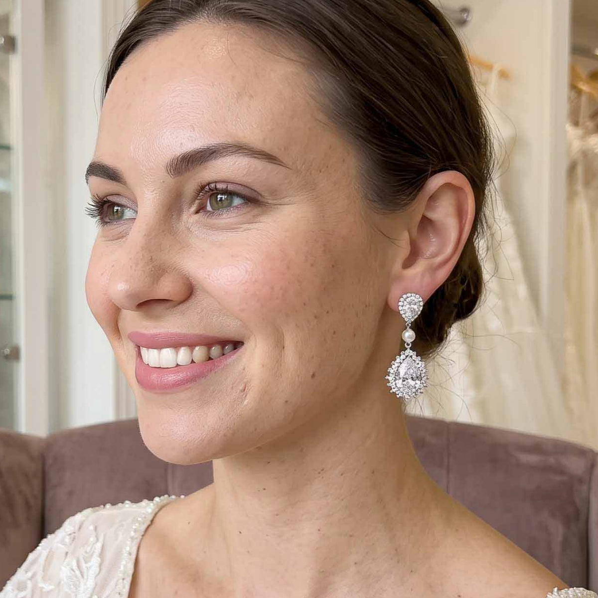 A smiling woman in profile wearing a sparkling Teardrop Cubic Zirconia and Pearl Bridal Earrings, seated on a mauve velvet chair in a bridal shop with white wedding dresses visible in the background.