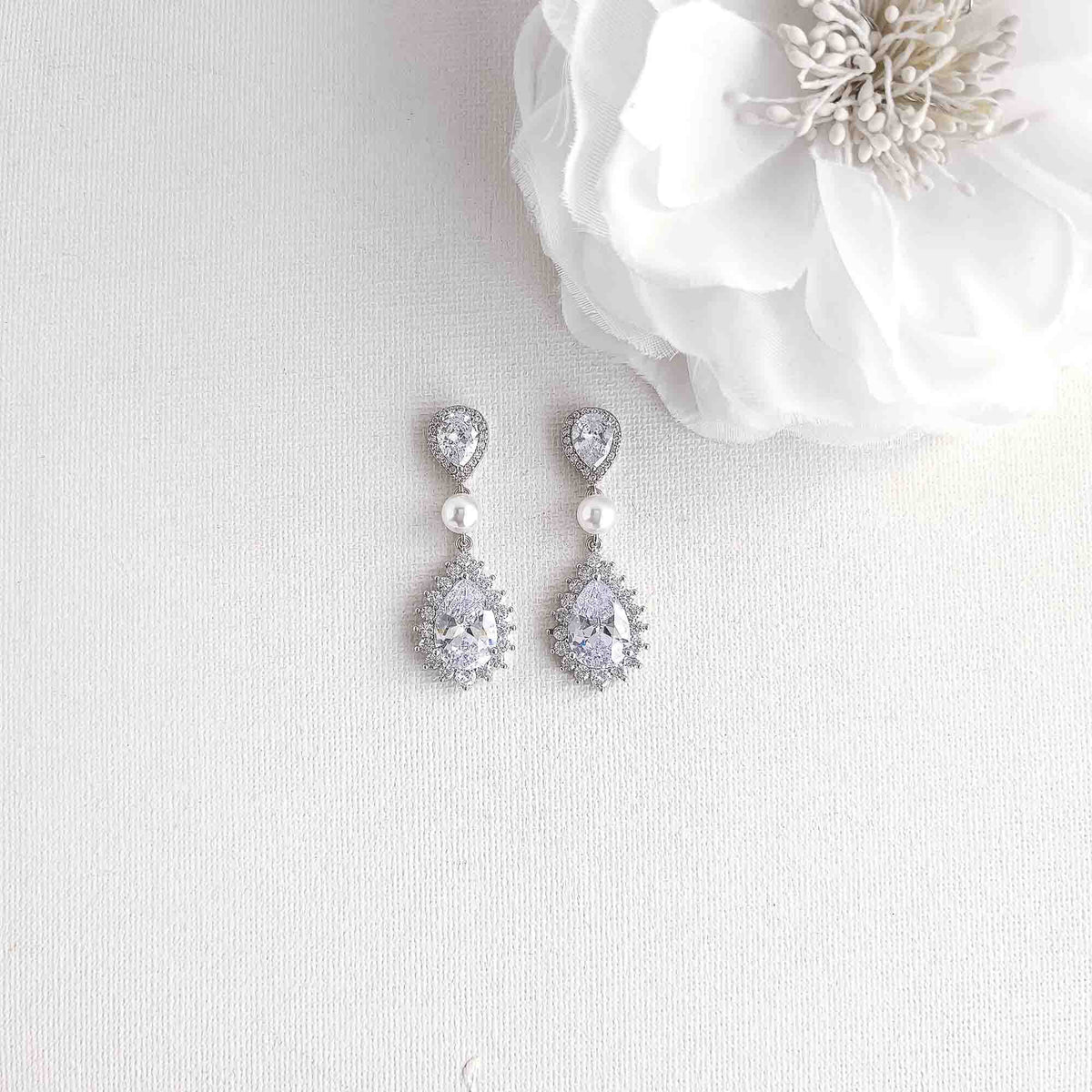 A pair of silver teardrop dangle earrings with clear stones and small pearl accents, displayed on a textured white fabric next to a white flower.
