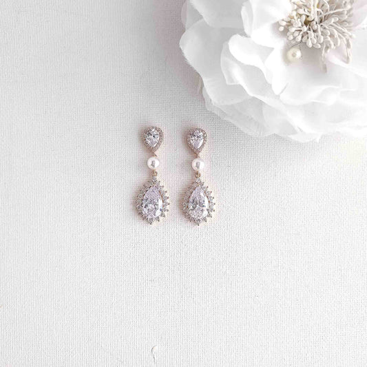A pair of rose gold drop earrings with clear teardrop crystals and small white pearls, displayed on a textured white fabric next to a white flower.