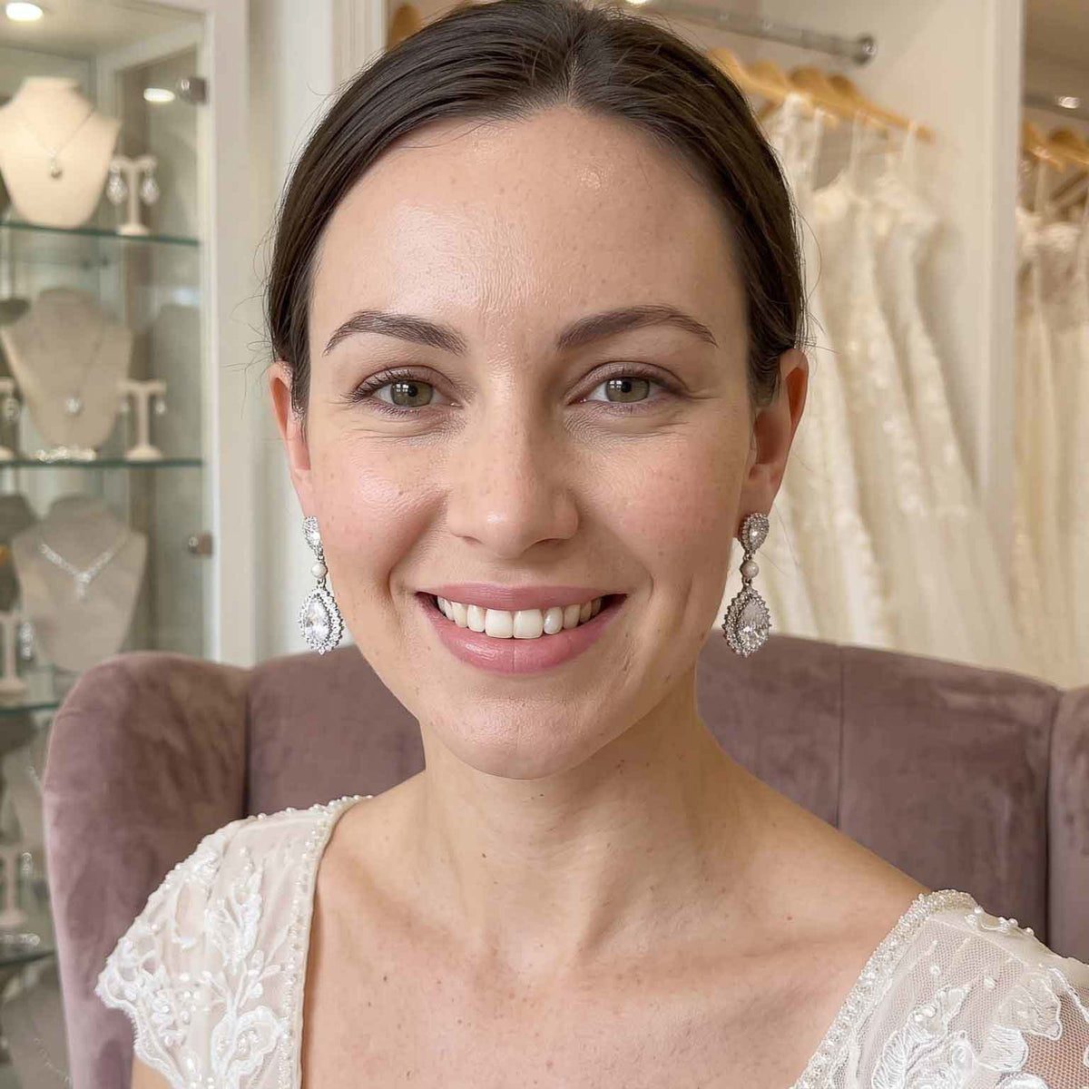 A smiling woman with dark hair pulled back wears sparkling silver teardrop dangle earrings and a white lace top, seated in a dusty mauve chair inside what appears to be a bridal or jewelry boutique.