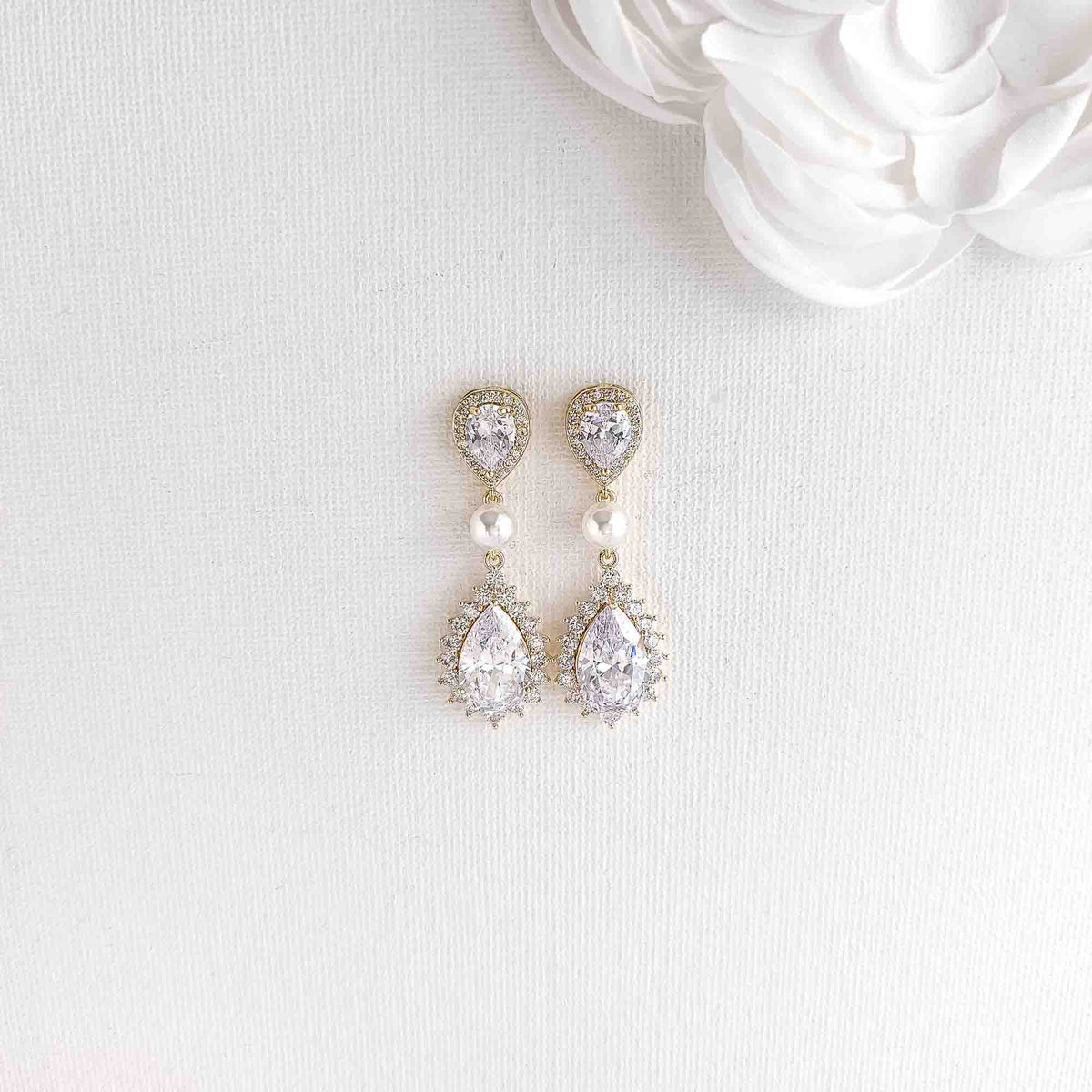 A pair of gold drop earrings with pear-cut clear crystals, small pearl beads, and halo accents, (Teardrop Cubic Zirconia and Pearl Bridal Earrings) displayed on a white textured fabric background with a white flower prop in the corner.