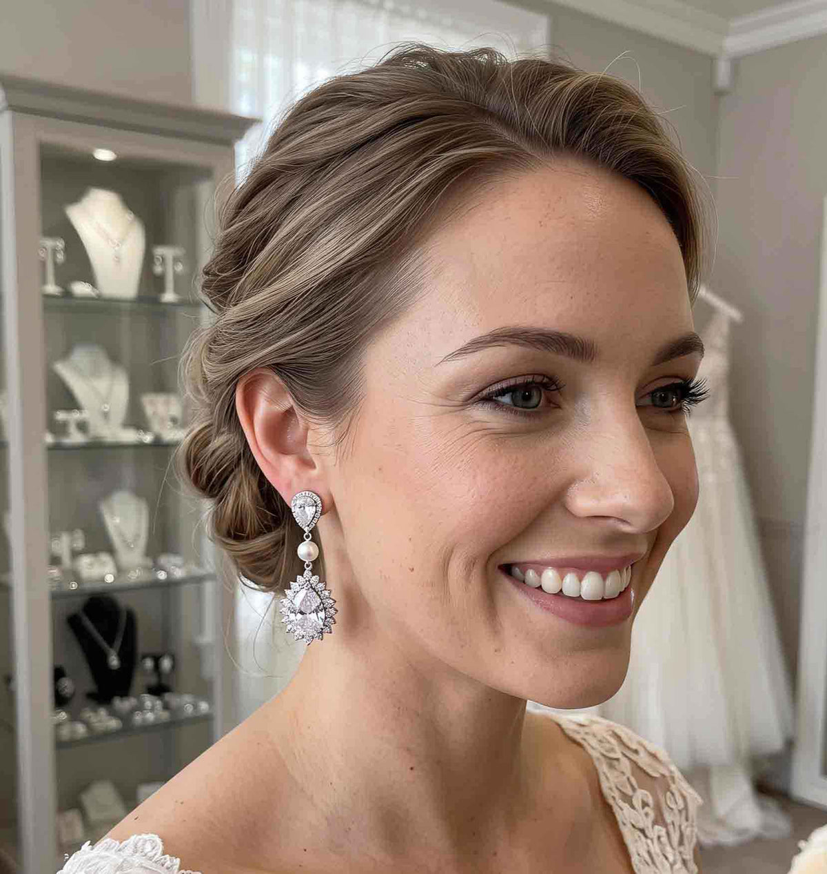 A smiling woman with styled brown hair wears a sparkling Teardrop Cubic Zirconia and Pearl Bridal Earrings. The photo is taken in a bridal boutique setting with jewelry display cases and a white dress visible in the background.