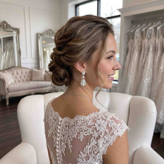 A woman in profile wearing a white lace bridal gown and dangling Teardrop Cubic Zirconia and Pearl Bridal Earrings, seated in a white armchair inside a bridal salon with mirrors, a sofa, and hanging wedding dresses in the background.